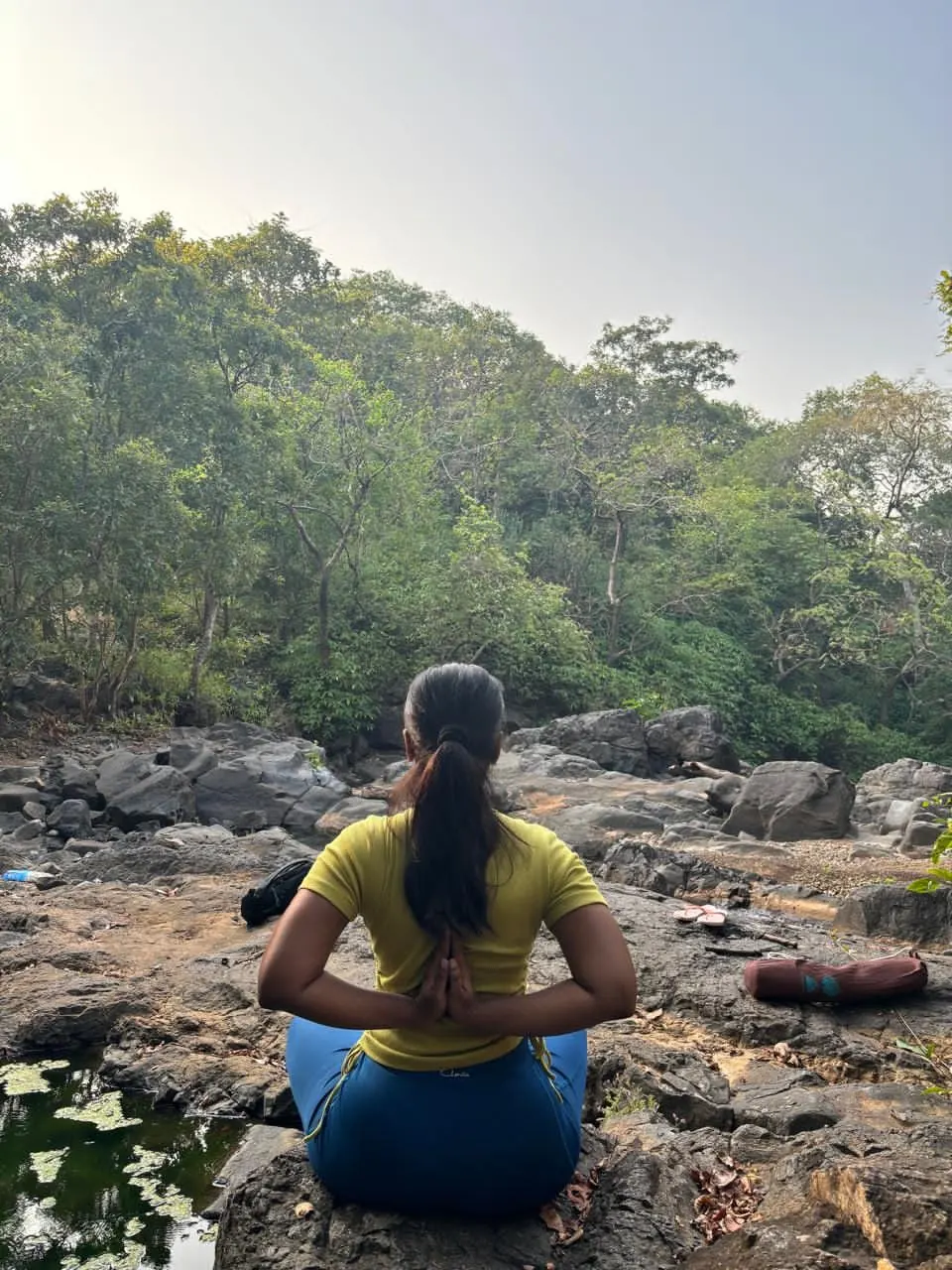 Neha sitting in reverse prayer pose, facing a green forest