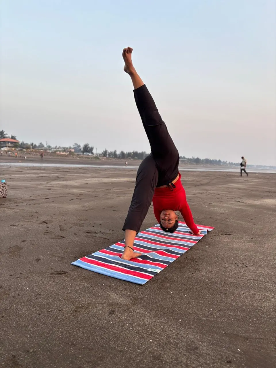 Neha in a standing split on a striped mat on the beach